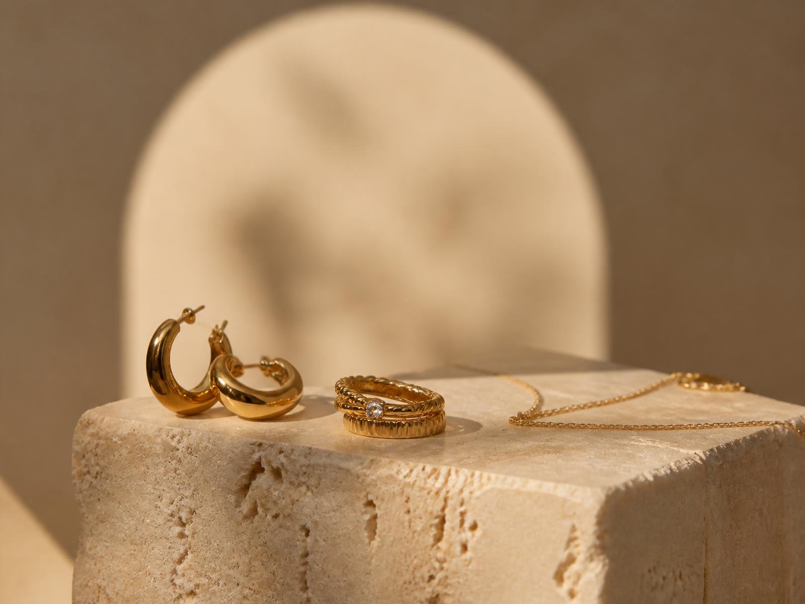 Gold hoop earrings, stacked rings and a fine chain on a travertine pedestal